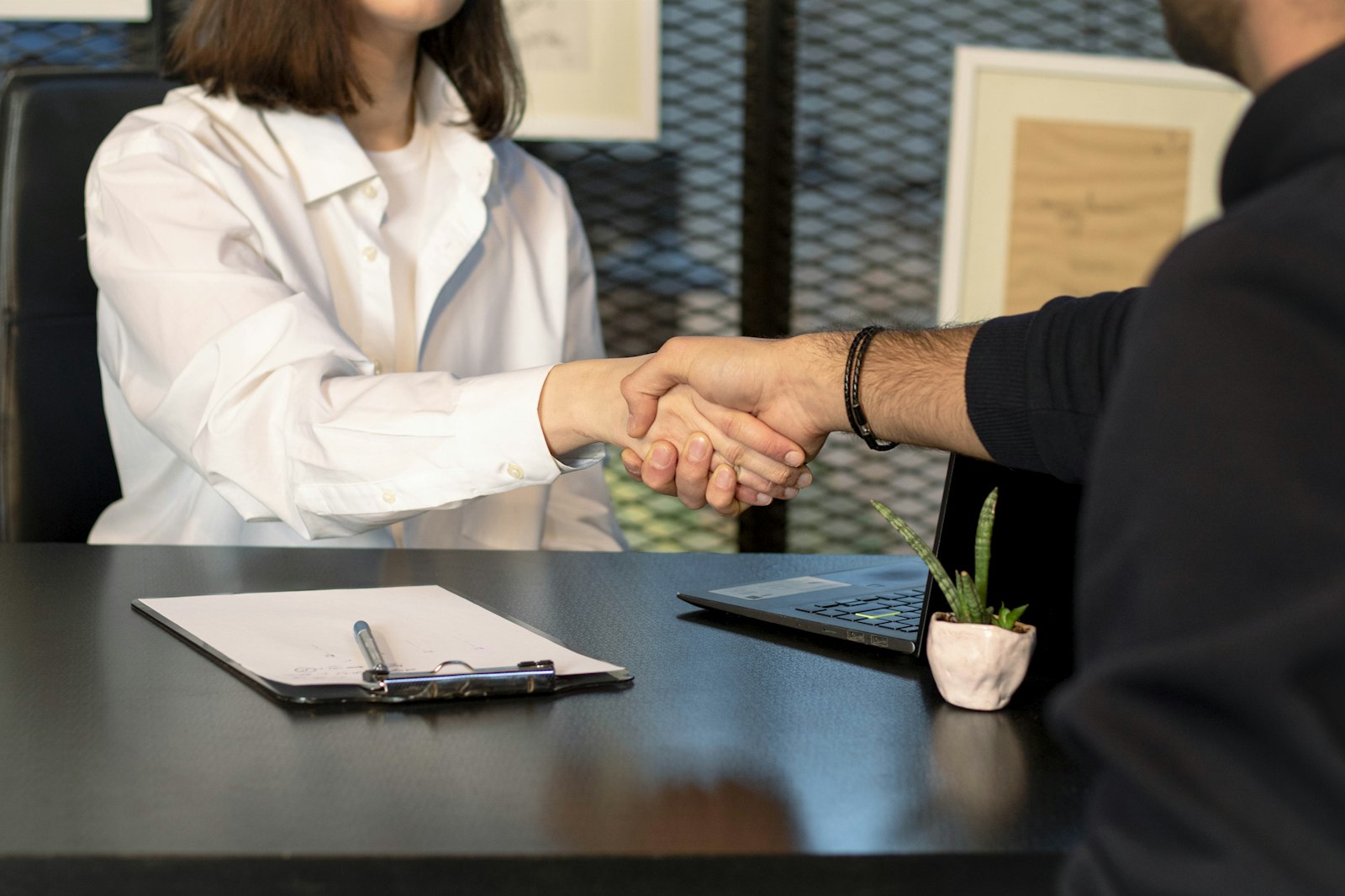 Two business people shaking hands over a laptop and clipboard, representing trust and partnership